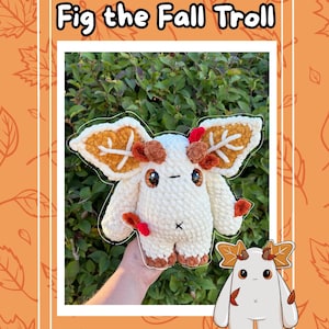 May include: A crocheted plush toy of a troll named "Fig the Fall Troll" is held in a hand. The troll is white with brown and orange accents, including leaf-shaped ears and feet. The image has an orange border with leaf designs.