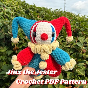 May include: A handmade crochet jester doll in red, blue, white, and yellow. The doll has a jester hat, a ruffled collar, and pom-poms. The text "Jinx the Jester Crochet PDF Pattern" is displayed on the image.
