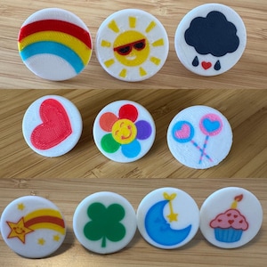May include: A collection of white, circular decorative buttons with colorful designs. The designs include a rainbow, a smiling sun with sunglasses, a rain cloud, a red heart, a flower, lollipops, a shooting star, a four-leaf clover, a moon and star, and a cupcake.