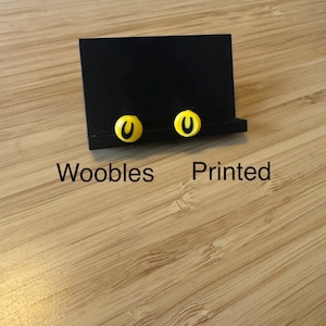 May include: A black display stand holding two yellow and black earrings. The earrings have a yellow base with a black "U" design. The text "Woobles Printed" is below the stand.