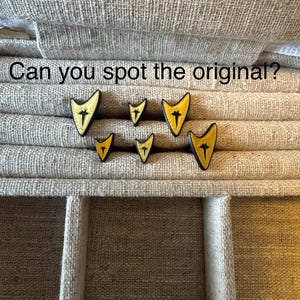 May include: A collection of yellow and black Star Trek insignia pins of varying sizes. The pins are arranged on a textured, neutral-toned surface. Text above the pins reads, "Can you spot the original?"