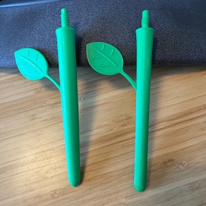 May include: Two bright green pens with leaf-shaped accents. The pens have a stem-like body and a leaf attached to the side. The pens are made of a solid material and are designed to resemble a plant.