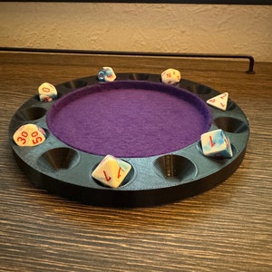 May include: A black circular dice tray with a purple felt lining and eight compartments. The tray has seven dice in it, including a d4, d6, d8, d10, d12, and two d20s. The dice are white with red numbers.
