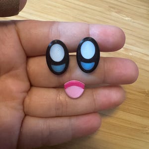 Könnte beinhalten: Ein Satz von drei Cartoon-Gesichtselementen: zwei schwarze ovale Augen mit weißen und blauen Highlights und ein rosa-roter lächelnder Mund. Die Elemente sind so angeordnet, dass sie ein Gesicht bilden.