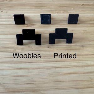May include: Two black plastic space invader figures, one with a smooth surface and the other with a textured surface. The words "Woobles" and "Printed" are written below the figures.