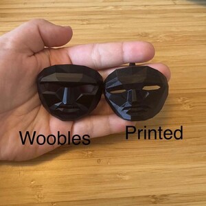 Puede incluir: Dos m&aacute;scaras negras de forma geom&eacute;trica se sostienen en una mano. La m&aacute;scara de la izquierda tiene la palabra "Woobles" impresa debajo. La m&aacute;scara de la derecha tiene la palabra "Printed" debajo. Las m&aacute;scaras tienen recortes para ojos y boca.