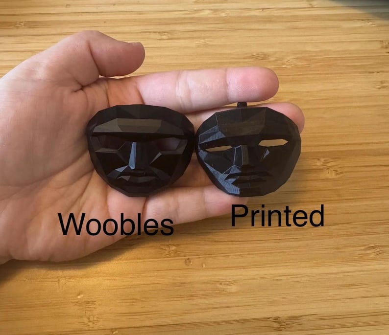 Puede incluir: Dos m&aacute;scaras negras de forma geom&eacute;trica se sostienen en una mano. La m&aacute;scara de la izquierda tiene la palabra "Woobles" impresa debajo. La m&aacute;scara de la derecha tiene la palabra "Printed" debajo. Las m&aacute;scaras tienen recortes para ojos y boca.