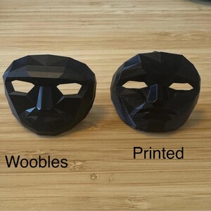 Puede incluir: Dos m&aacute;scaras negras de forma geom&eacute;trica con aberturas para los ojos y la boca. Una m&aacute;scara est&aacute; etiquetada como "Woobles" y la otra como "Printed". Las m&aacute;scaras se muestran sobre una superficie de madera marr&oacute;n claro.