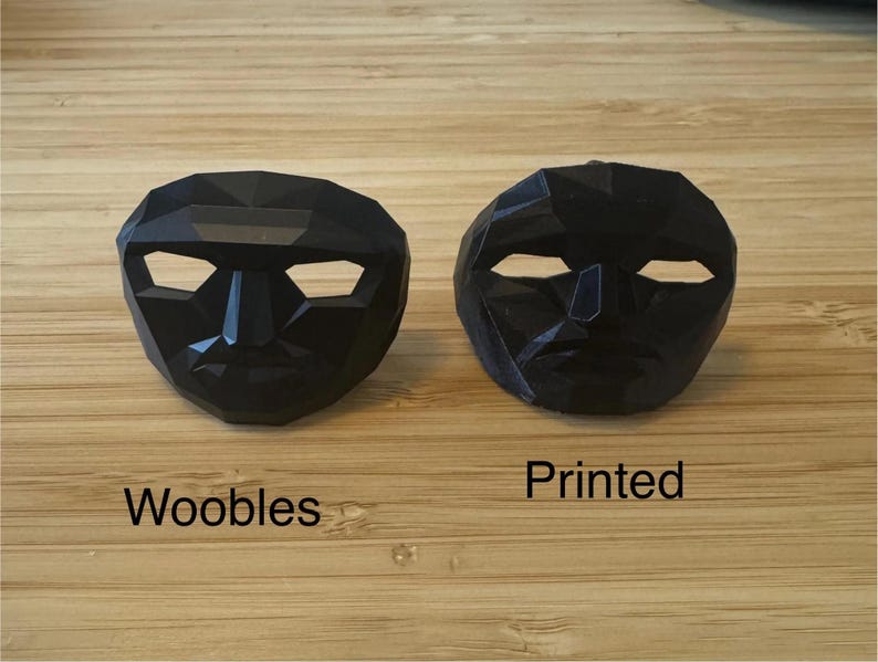 Puede incluir: Dos m&aacute;scaras negras de forma geom&eacute;trica con aberturas para los ojos y la boca. Una m&aacute;scara est&aacute; etiquetada como "Woobles" y la otra como "Printed". Las m&aacute;scaras se muestran sobre una superficie de madera marr&oacute;n claro.