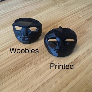 Puede incluir: Dos m&aacute;scaras faciales negras y geom&eacute;tricas. Una m&aacute;scara tiene la palabra "Woobles" impresa debajo. La otra m&aacute;scara tiene la palabra "Printed" impresa debajo. Las m&aacute;scaras est&aacute;n sobre una superficie de madera marr&oacute;n claro.