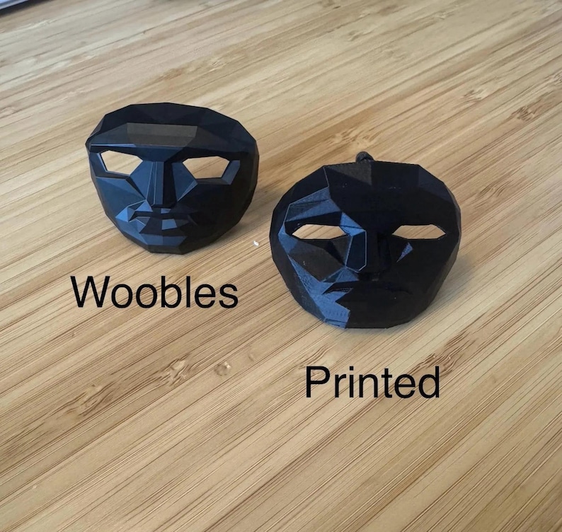 Puede incluir: Dos m&aacute;scaras faciales negras y geom&eacute;tricas. Una m&aacute;scara tiene la palabra "Woobles" impresa debajo. La otra m&aacute;scara tiene la palabra "Printed" impresa debajo. Las m&aacute;scaras est&aacute;n sobre una superficie de madera marr&oacute;n claro.