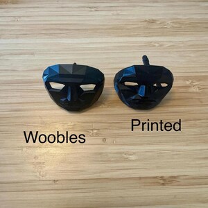 Puede incluir: Dos m&aacute;scaras faciales negras y geom&eacute;tricas con ranuras para los ojos blancas sobre una superficie de madera. Una m&aacute;scara est&aacute; etiquetada como "Woobles", y la otra como "Printed".