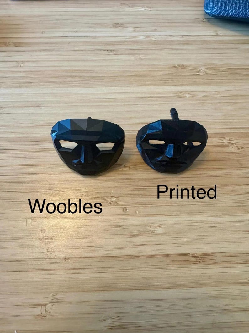 Puede incluir: Dos m&aacute;scaras faciales negras y geom&eacute;tricas con ranuras para los ojos blancas sobre una superficie de madera. Una m&aacute;scara est&aacute; etiquetada como "Woobles", y la otra como "Printed".