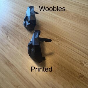 Puede incluir: Dos objetos negros de forma geom&eacute;trica con un tallo, etiquetados como "Woobles" y "Printed" sobre una superficie de madera. Los objetos tienen un dise&ntilde;o facetado y parecen estar impresos en 3D.