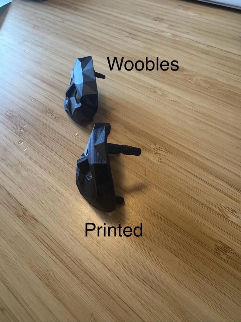 Puede incluir: Dos objetos negros de forma geom&eacute;trica con un tallo, etiquetados como "Woobles" y "Printed" sobre una superficie de madera. Los objetos tienen un dise&ntilde;o facetado y parecen estar impresos en 3D.