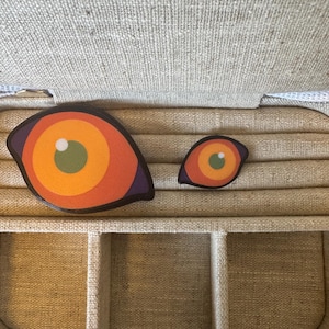 May include: Two eye-shaped decorative pieces with orange, yellow, and green concentric circles, and a purple border. They are placed on a beige fabric surface, possibly inside a jewellery box or storage container.
