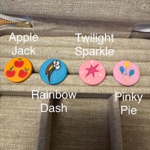 Op de afbeelding: Vier platte, ronde bedels met symbolen van My Little Pony-personages. De oranje bedel toont drie rode appels, de blauwe heeft een regenboogwolk, de roze toont een magentakleurige ster en de lichtroze bedel toont drie ballonnen.