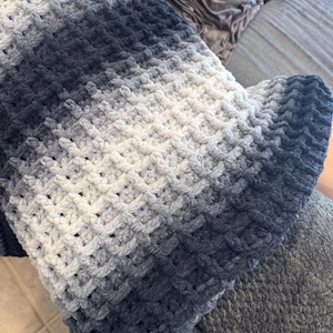 Peut inclure: Une couverture douce, crochetée à la main, dans un dégradé de gris et de bleu marine. La couverture présente un motif texturé et tissé et est pliée sur une surface grise, mettant en valeur la transition de couleur de clair à foncé.