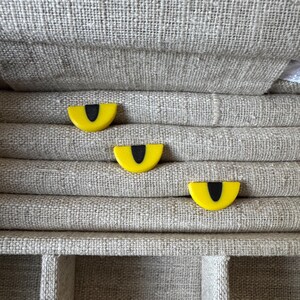 May include: Three small, semi-circular stud earrings featuring a bright yellow background with a vertical black slit pupil design, resembling cat eyes. Each piece has a smooth, flat surface and a distinct, curved shape that mimics an animal eye.