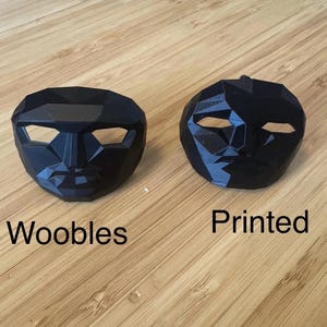 Puede incluir: Dos m&aacute;scaras geom&eacute;tricas negras se muestran sobre una superficie de madera. Una m&aacute;scara est&aacute; etiquetada como "Woobles", y la otra como "Printed". Las m&aacute;scaras tienen aberturas para los ojos y un dise&ntilde;o de estructura facial.