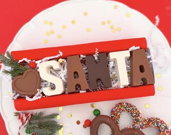 May include: A red box with chocolate letters spelling "SANTA" on a white plate. The "S" is white, the "A" is brown, the "N" is dark brown, the "T" is white, and the final "A" is brown. A chocolate heart and pretzel are also visible.