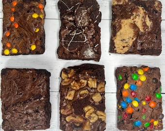 Peut inclure: Six brownies assortis avec diverses garnitures, dont des bonbons au chocolat, des noix et des biscuits. Les brownies rectangulaires sont disposés sur une surface en bois blanc. Les garnitures ajoutent de la couleur et de la texture aux pâtisseries brun foncé.