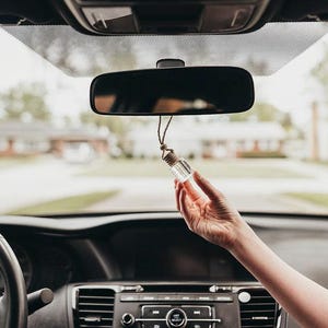 May include: A clear glass bottle with a brown string hanging from the rear-view mirror of a car. The bottle is filled with a clear liquid and has a cork stopper.