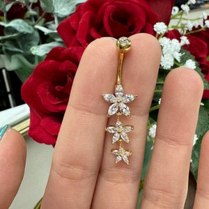 May include: Gold body jewellery with a dangling chain of three flower-shaped charms, each adorned with clear cubic zirconia stones.