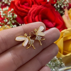 May include: A pair of gold-colored fairy stud earrings with white enamel wings and rhinestone accents. The earrings are held in a hand, with a blurred background of red and yellow roses.