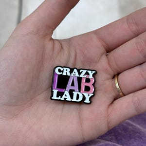 Puede incluir: Un pin rectangular esmaltado con las palabras "CRAZY LAB LADY" en blanco, negro y rosa. El pin se sostiene en una mano con un anillo de oro en el dedo anular. El fondo es una tela morada.