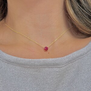 May include: A delicate gold chain necklace featuring a single, round, vibrant red gemstone pendant set in a simple four-prong setting. The necklace rests against the neck, showcasing the thin metallic links and the bright, faceted red stone.