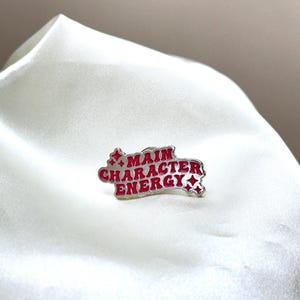 Puede incluir: Un pin de esmalte plateado con una inscripción roja que dice "MAIN CHARACTER ENERGY" con estrellas a cada lado.