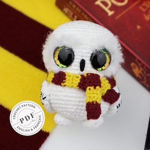 Può includere: Gufo bianco all'uncinetto con una sciarpa a righe rosse e gialle. Il gufo ha grandi occhi scintillanti. Il gufo è seduto su una superficie bianca con una coperta a righe rosse e gialle e un libro sullo sfondo.