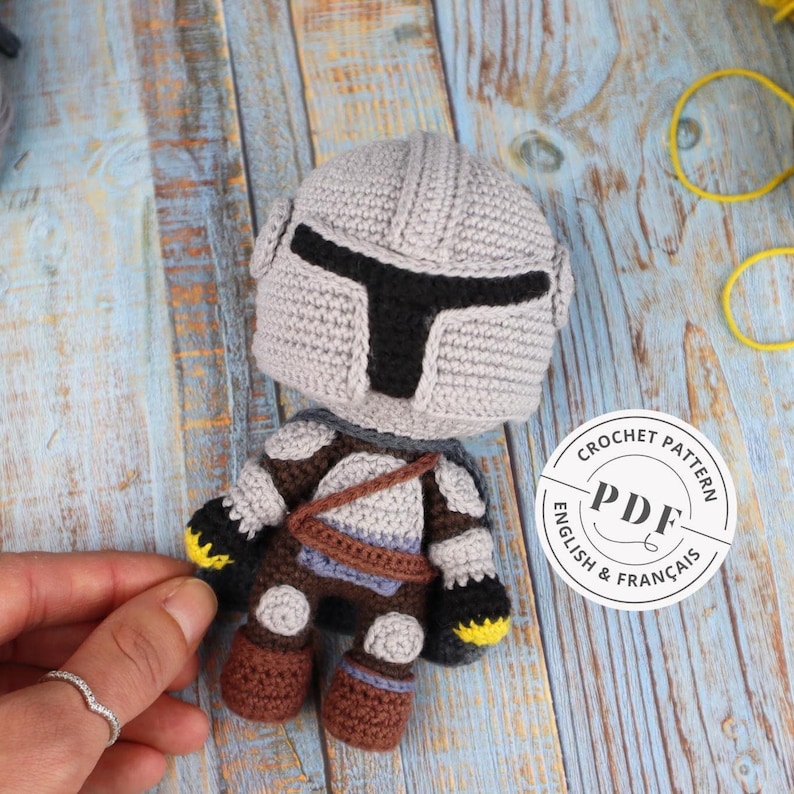 K&ouml;nnte beinhalten: H&auml;kelanleitung f&uuml;r eine graue und braune Amigurumi-Figur mit einem Helm mit schwarzem Visier. Die Anleitung ist in Englisch und Franz&ouml;sisch verf&uuml;gbar. Die Anleitung ist ein PDF-Download.