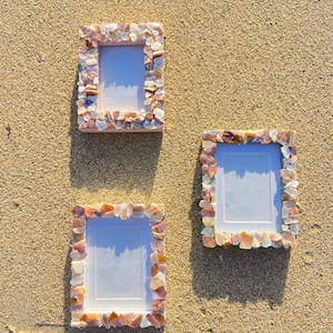 May include: Three rectangular photo frames decorated with small, colorful seashells. The frames are arranged on a sandy surface, with the shells in shades of pink, beige, and white. The frames have a white interior.
