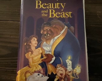 Puede incluir: Una caja de cinta VHS con la ilustración de la portada de la película clásica de Disney "La Bella y la Bestia". Se puede ver a Bella y la Bestia bailando, con otros personajes de la película, incluyendo Lumière, Din Don y la Señora Tetera, en el fondo.