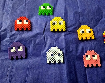 Può includere: Una collezione di figure in pixel art realizzate con piccole perline di plastica, tra cui un personaggio circolare giallo con la bocca aperta e diverse figure a forma di fantasma nei colori rosso, verde, giallo, viola, bianco, rosa e arancione.