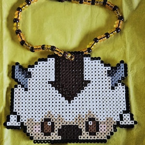 May include: A necklace featuring a pendant made of white, brown, and grey plastic fuse beads arranged in the shape of a cartoon creature with a brown arrow on its head. The pendant is attached to a circular strand of alternating brown and gold beads.