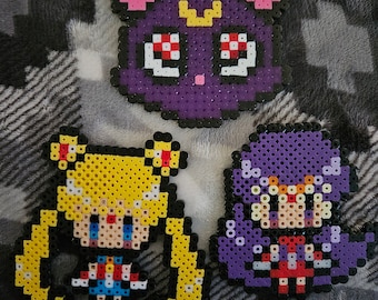 May include: Three pixel art figures made of colorful beads. The figures are of Sailor Moon, Luna, and Sailor Mercury. Sailor Moon is wearing a yellow dress with a blue and red skirt. Luna is a purple cat with pink ears and yellow eyes. Sailor Mercury is wearing a purple dress with a red and white skirt.