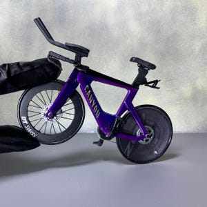 May include: A miniature purple Canyon road bike with black wheels and components. The bike features the Canyon logo in white, DT Swiss branding on the front wheel, and a sleek, aerodynamic design. The bike is held by a gloved hand.