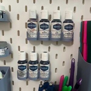 May include: A collection of AmeriColor gel food color bottles in various shades of red, brown, and blue, including Tulip Red, Maroon, Dijon, Warm Brown, Olive, Navy Blue, and Super Black. The bottles are displayed on a white pegboard with other craft supplies.