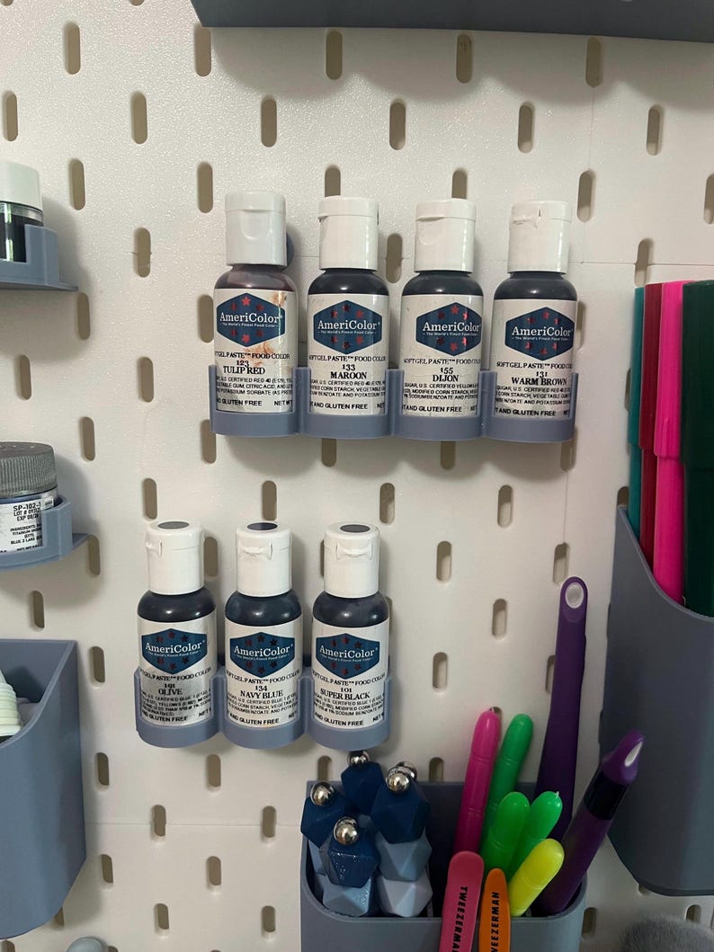 May include: A collection of AmeriColor gel food color bottles in various shades of red, brown, and blue, including Tulip Red, Maroon, Dijon, Warm Brown, Olive, Navy Blue, and Super Black. The bottles are displayed on a white pegboard with other craft supplies.
