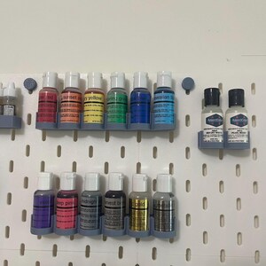 May include: A collection of food coloring bottles in various colors, including red, orange, yellow, green, blue, and purple. The bottles are arranged on a white pegboard, with labels indicating the color names. The image showcases a range of vibrant hues.