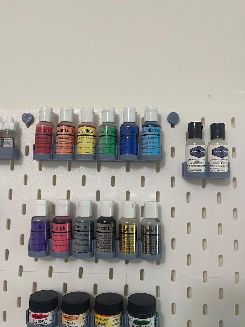 May include: A collection of food coloring bottles in various colors, including red, orange, yellow, green, blue, and purple. The bottles are arranged on a white pegboard, with labels indicating the color names. The image showcases a range of vibrant hues.