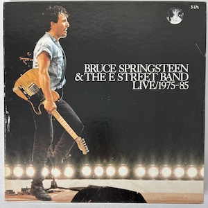 May include: A black and white album cover featuring Bruce Springsteen and the E Street Band. The cover shows Springsteen standing on a stage with a guitar in his hand. The text on the cover reads "Bruce Springsteen & The E Street Band LIVE/1975-85".