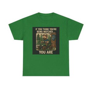 May include: Green t-shirt with a graphic that reads "IF YOU THINK YOU'RE BEING WATCHED... YOU ARE." The graphic depicts a person on an ATV in a forest setting, with a tree and a creature with blue skin.