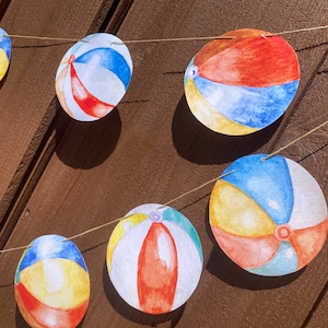 Peut inclure: Une guirlande de découpes de papier de ballons de plage colorés, chacun avec un design différent, est suspendue sur un fond de bois brun. Les ballons de plage sont peints dans des tons de bleu, rouge, jaune et blanc.