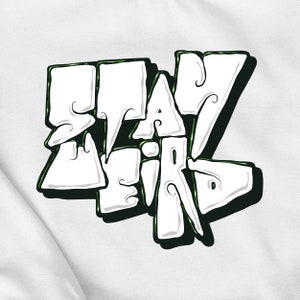 Könnte beinhalten: Weißes Grafikdesign mit schwarzen Umrissen und grünen Akzenten. Das Design sagt "Stay Weird" in einer stilisierten Schrift.