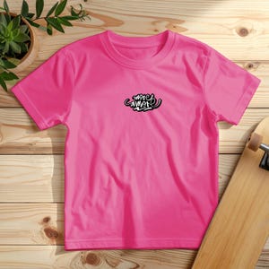 Könnte beinhalten: Ein leuchtend pinkfarbenes T-Shirt mit Rundhalsausschnitt. Das Shirt zeigt die Worte "more amor" in einem schwarz-weißen Schriftzug-Design auf der oberen Brust. Das T-Shirt wird auf einer Holzoberfläche präsentiert.