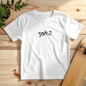 Könnte beinhalten: Weißes T-Shirt mit dem Wort "yeah" in schwarzer Schrift. Das T-Shirt hat einen Rundhalsausschnitt und besteht aus einem weichen Material. Das T-Shirt liegt auf einer Holzoberfläche.